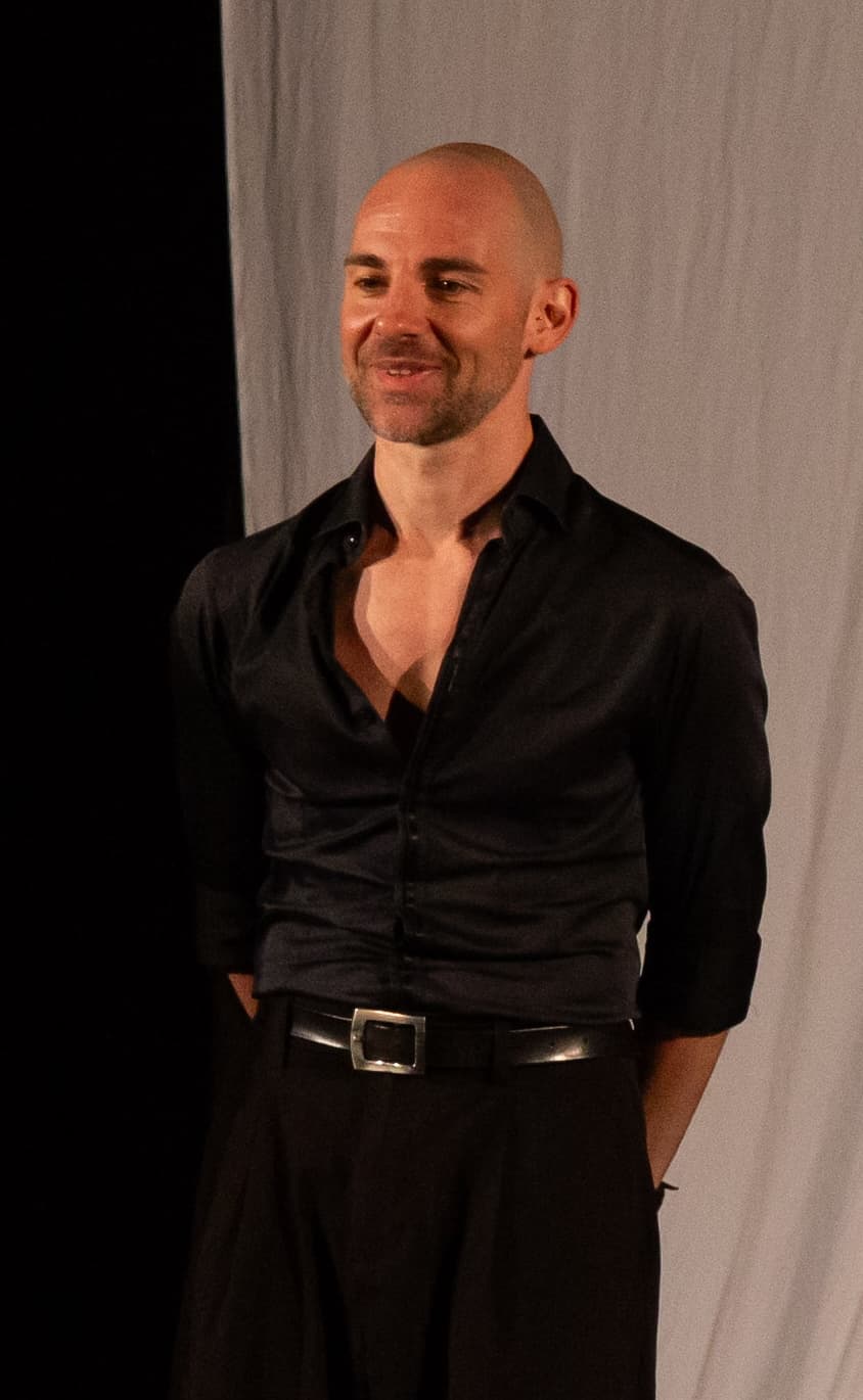 Uomo calvo sorridente con camicia nera sbottonata e pantaloni scuri davanti a una tenda.