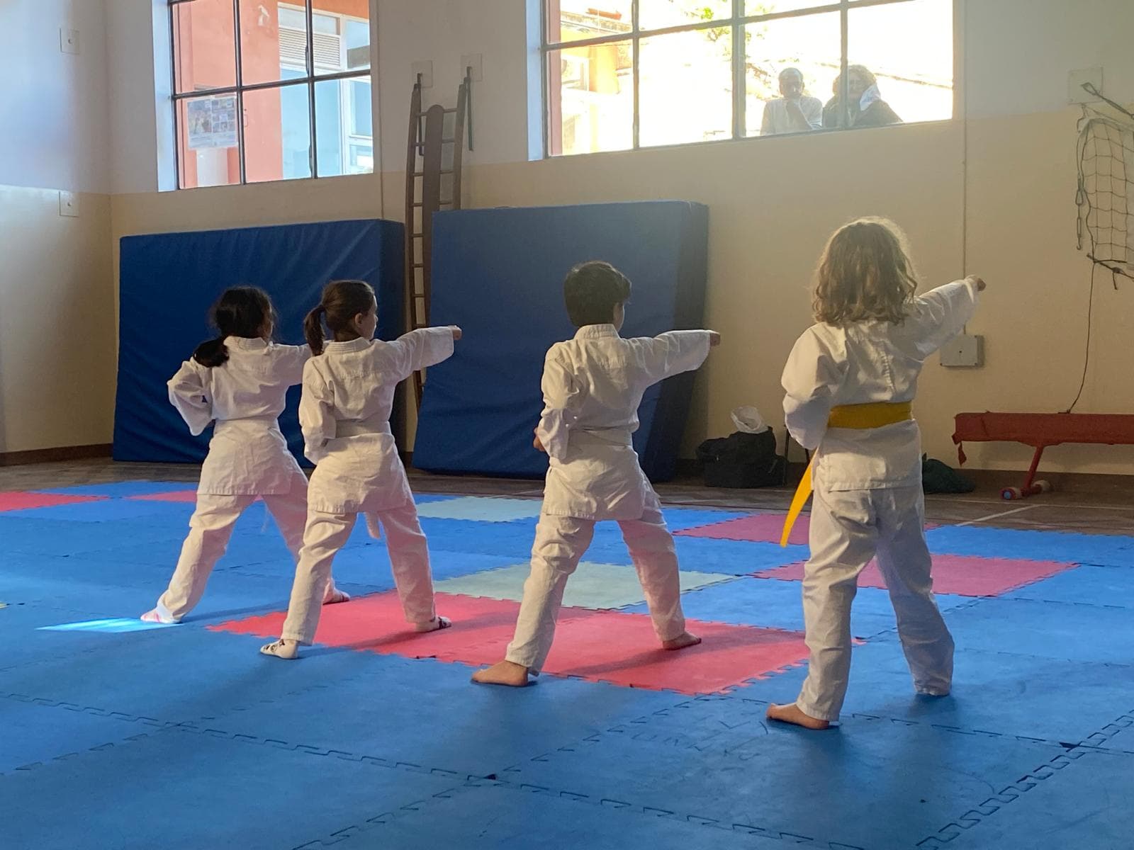 Quattro bambini in divisa bianca praticano karate su un tatami blu e rosso in palestra.