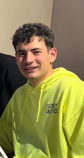 Uomo sorridente con capelli ricci scuri e felpa giallo neon del Genova Sport College.