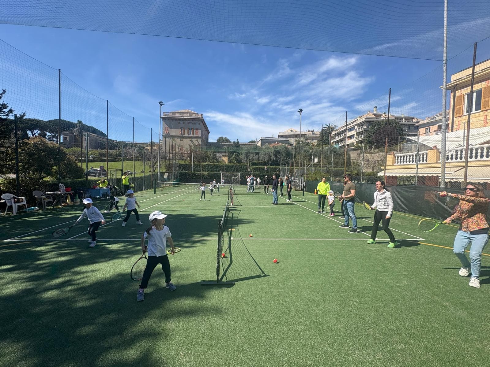 Bambini e adulti giocano a tennis su un campo verde all'aperto in una giornata soleggiata.