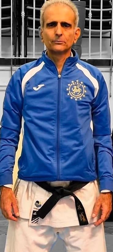 Uomo con capelli grigi in giacca blu e divisa da karate con cintura nera.