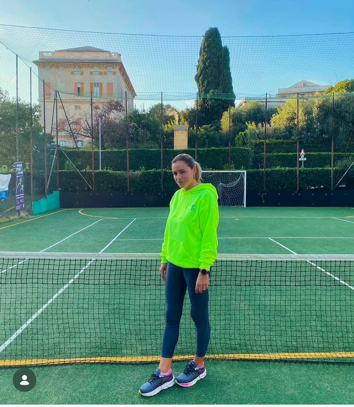 Donna in felpa giallo fluo e leggings blu dietro la rete di un campo sportivo.