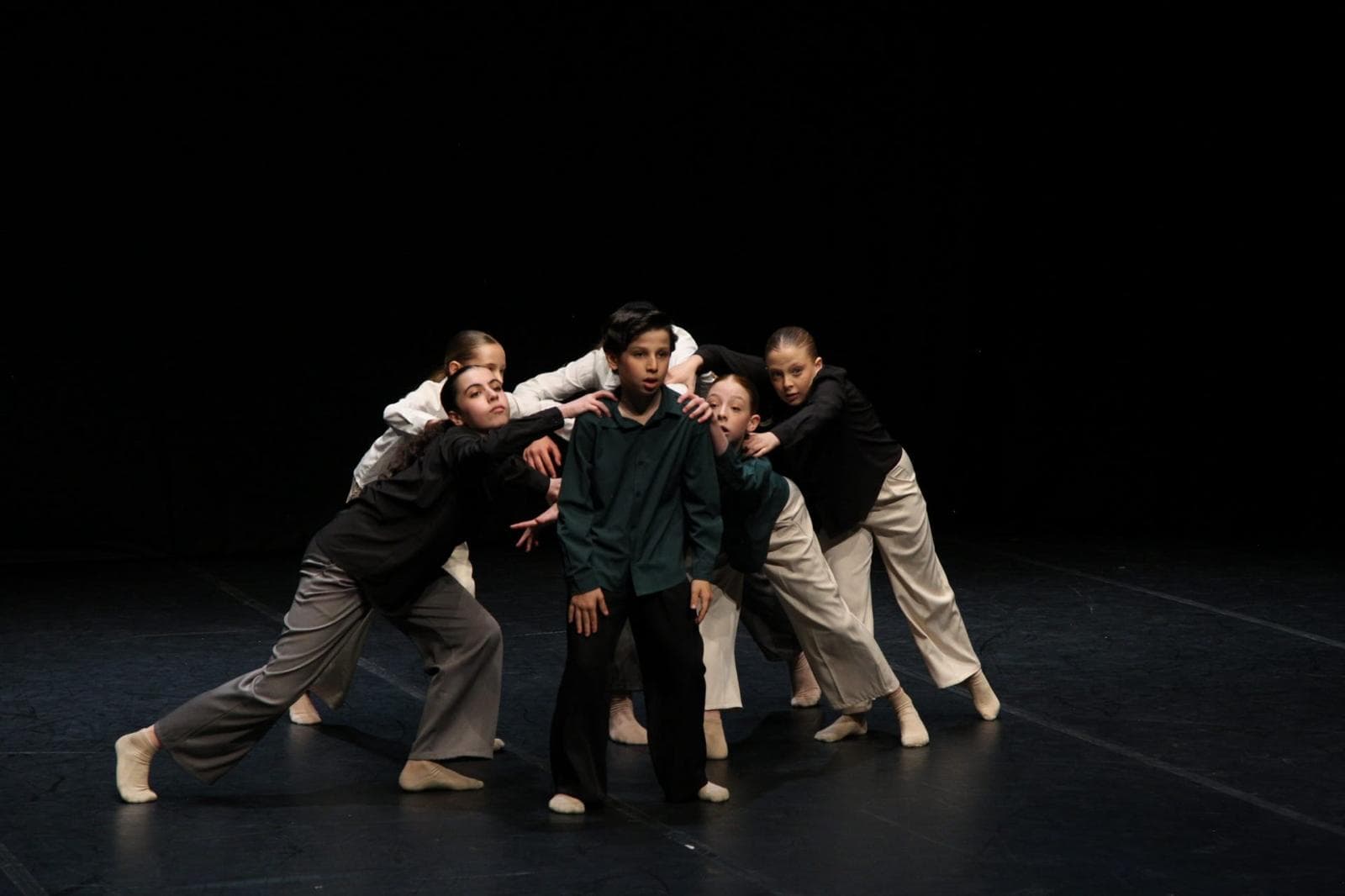 Giovani ballerini in scena circondano un ragazzo con camicia verde in una coreografia drammatica.