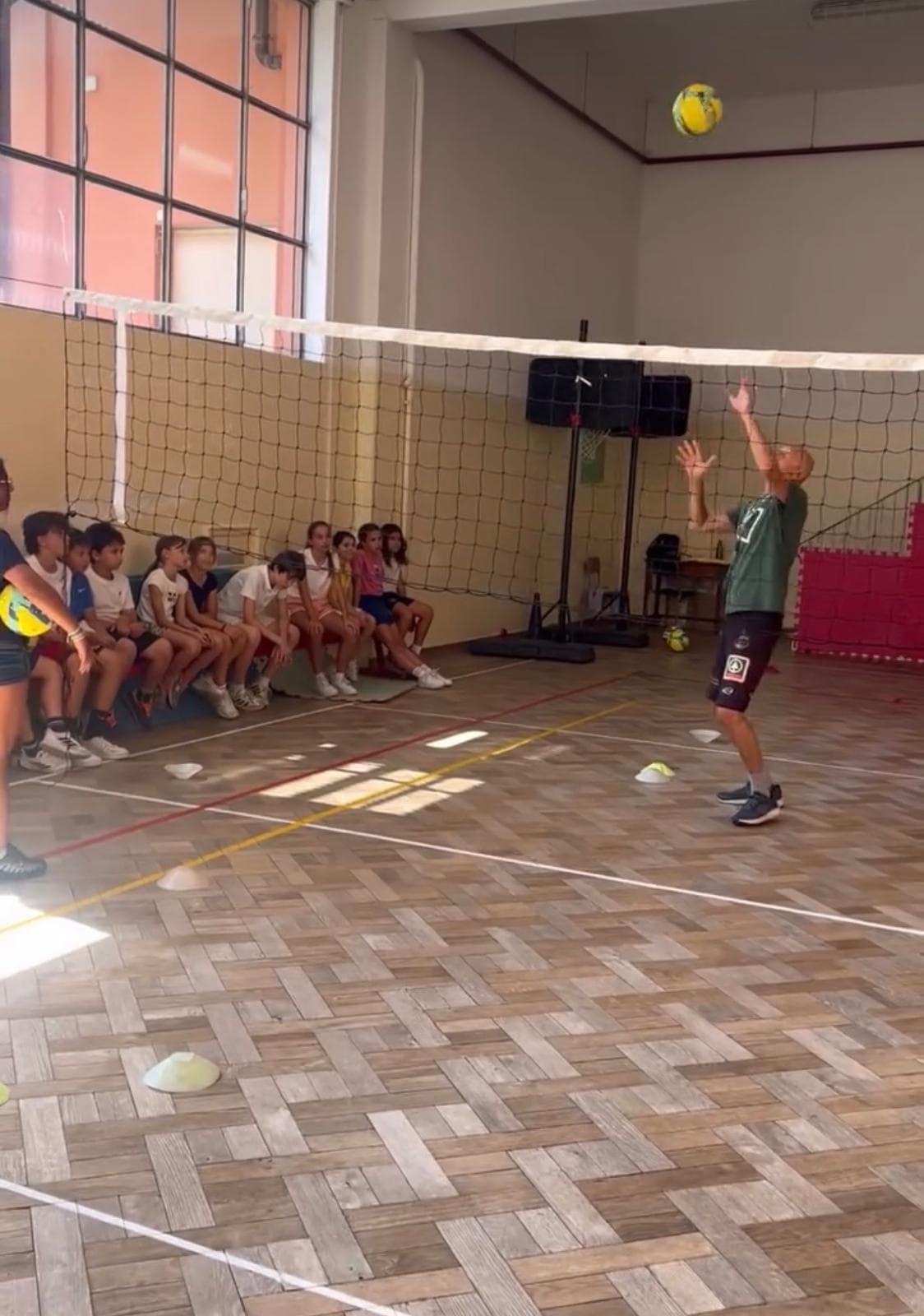 Uomo colpisce un pallone da pallavolo in palestra mentre dei bambini osservano dalla panchina.
