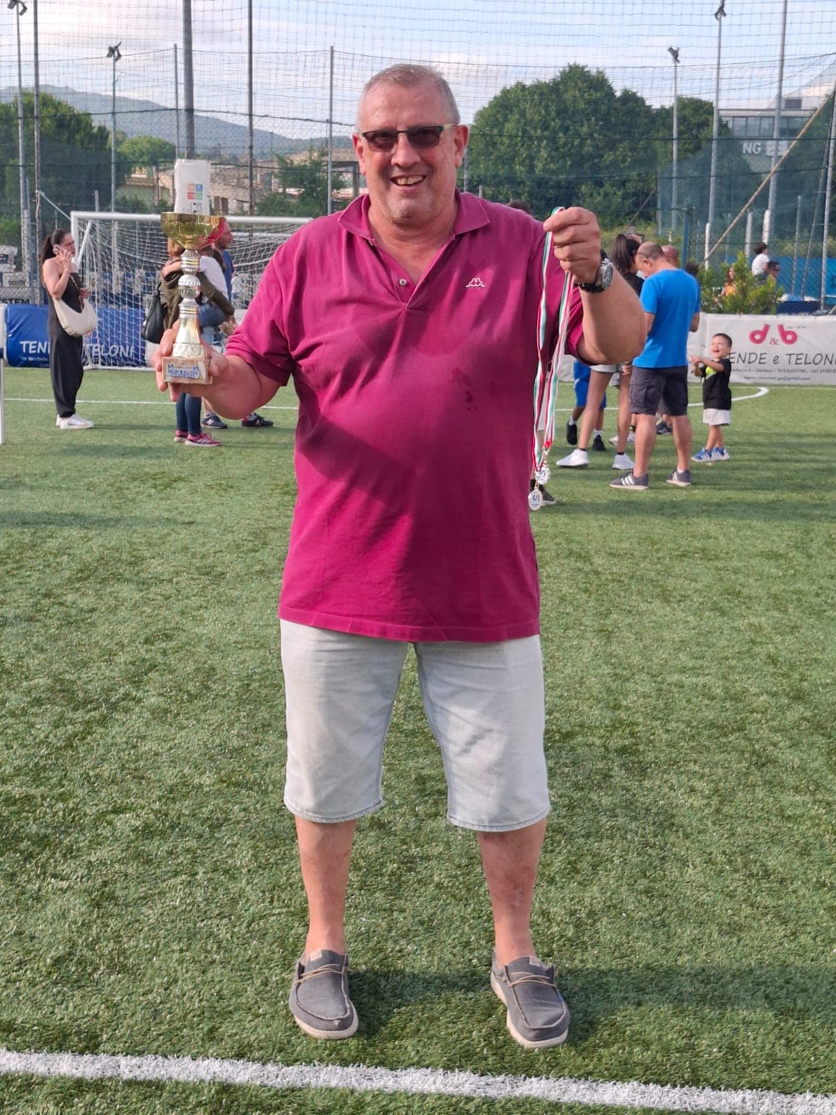 Uomo sorridente in polo fucsia con trofeo e medaglia su un campo da calcio.