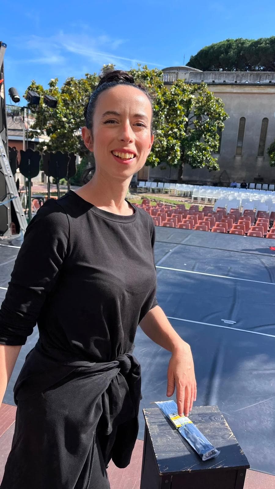 Donna sorridente in abito nero su un palco all'aperto con sedie vuote sullo sfondo.