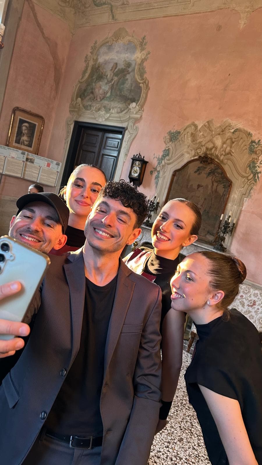 Cinque persone sorridenti scattano un selfie in una sala elegante decorata con affreschi classici.