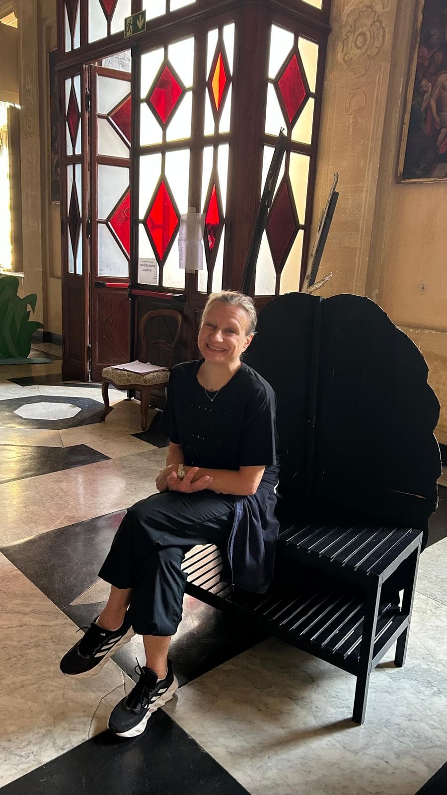 Donna sorridente seduta su una sedia nera a forma di conchiglia in un ambiente storico.