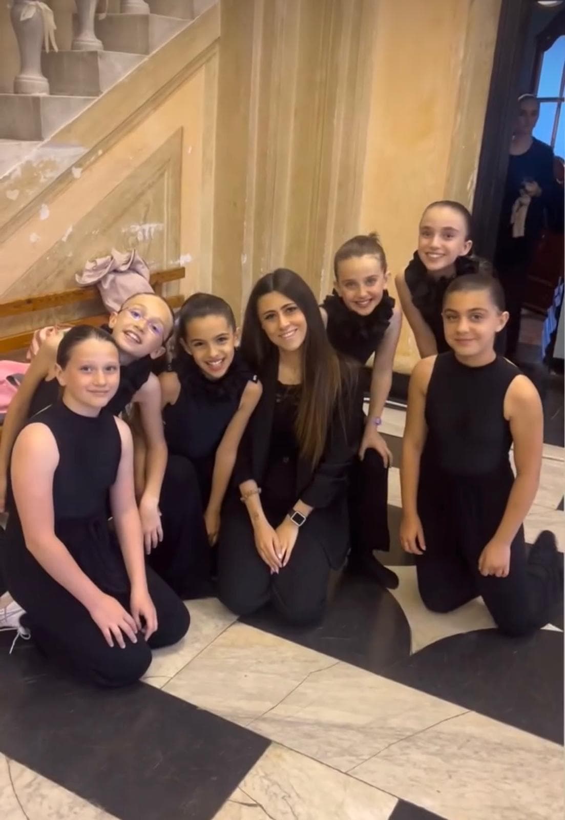Gruppo di giovani ballerine in nero sorride con l'insegnante su un pavimento a scacchi.