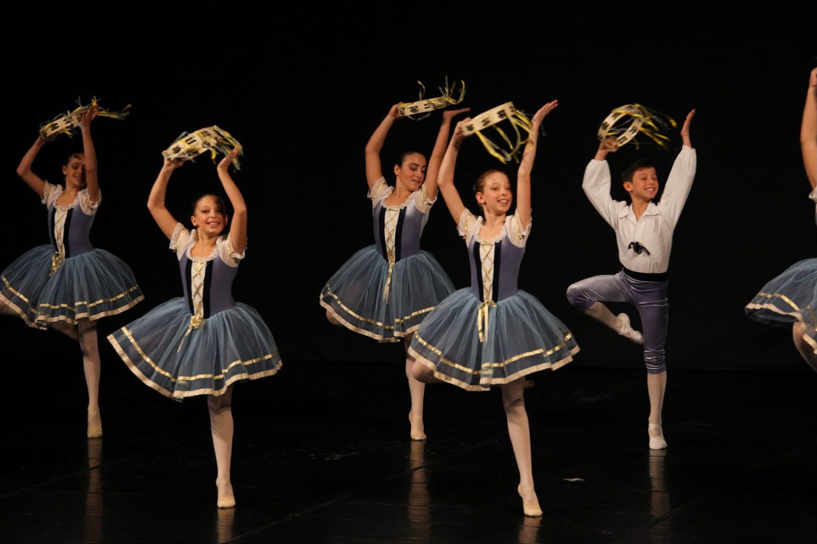 Ballerini classici in tutù azzurri e bianchi sollevano tamburelli durante una coreografia su sfondo nero.