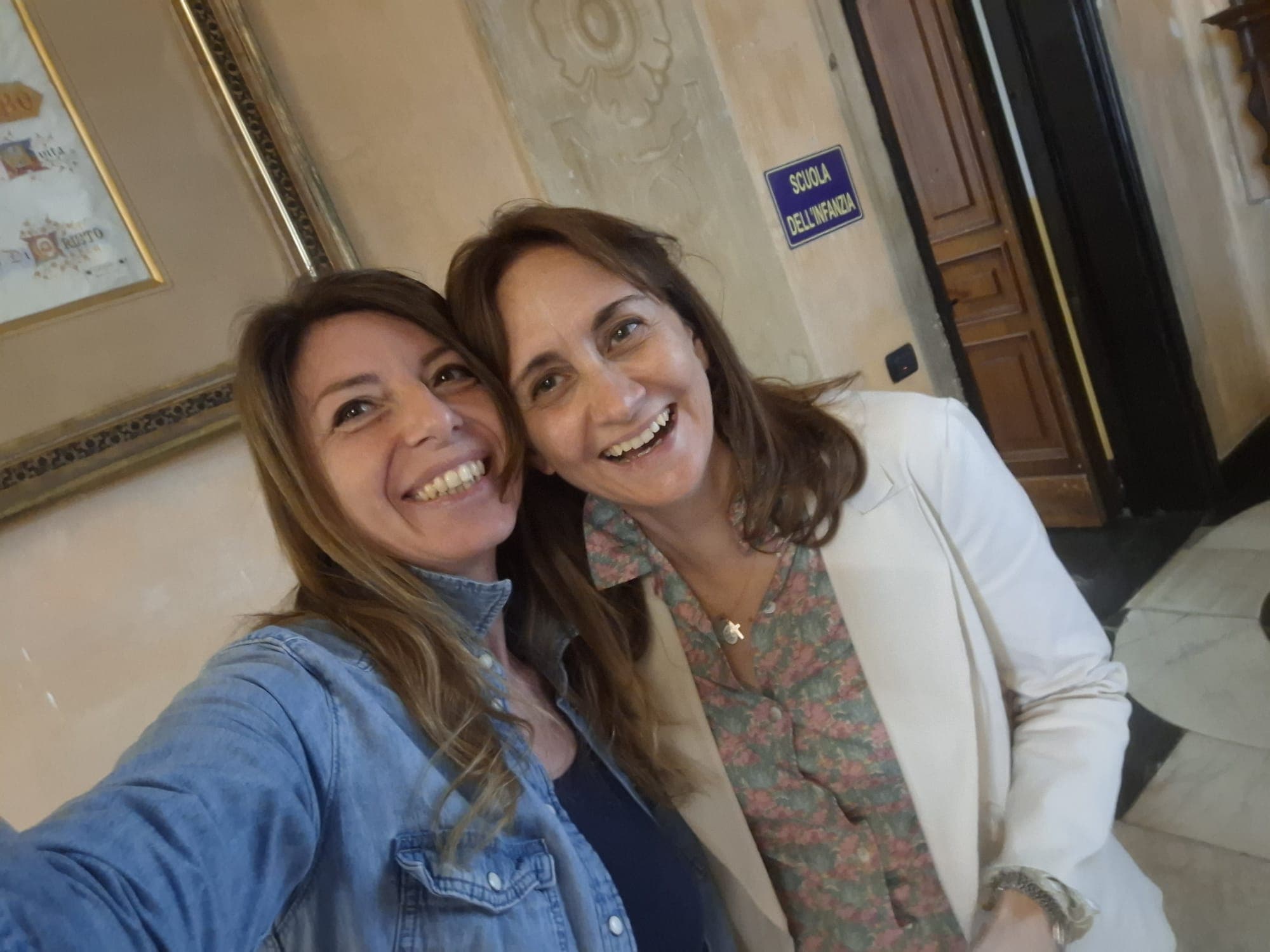 Due donne sorridenti scattano un selfie vicino a un cartello della scuola dell'infanzia.