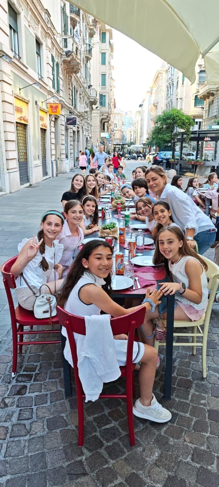 Bambini e adulti sorridenti seduti a una lunga tavola all'aperto in una via acciottolata.