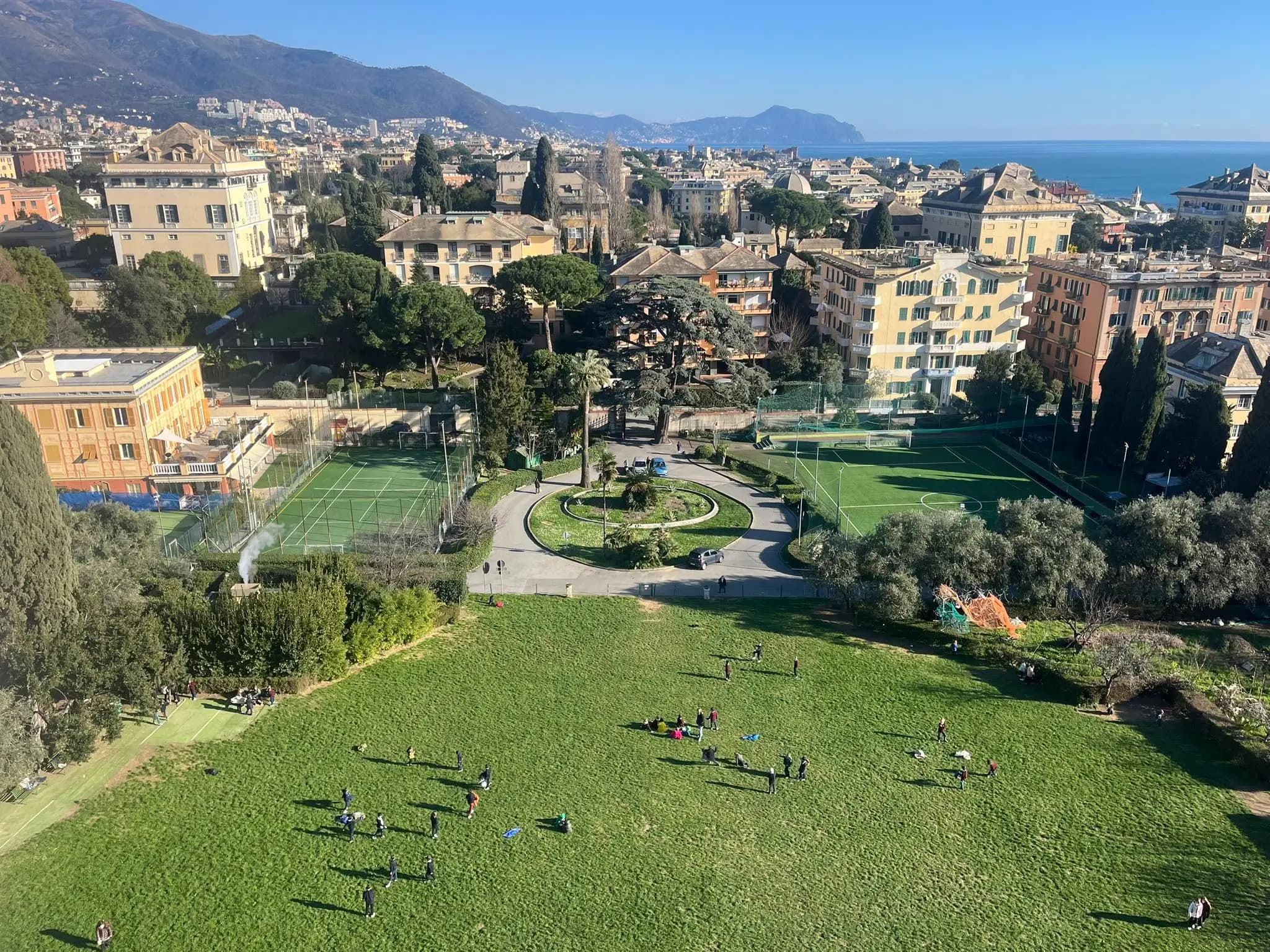 Veduta aerea di un parco con campi sportivi, edifici storici e mare sullo sfondo.